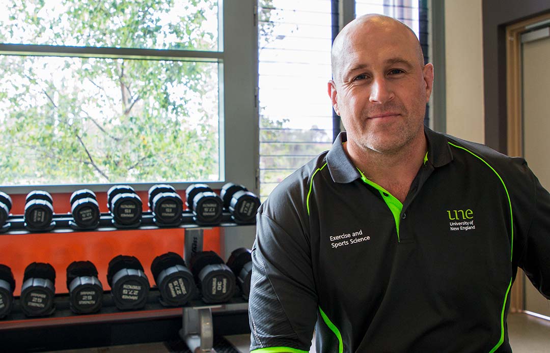 UNE's new lecturer in Exercise and Sports Science Bradley Morris smiles at the camera with a set of weights in the background.