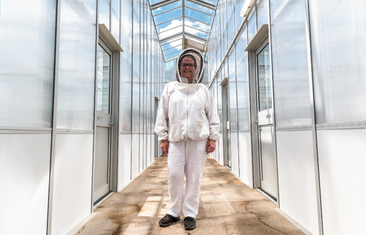 A woman stands in a light-filled corridor with a glass roof. She wears beekeeping protective clothing of white overalls and a hood with netting over the face. She wears black-rimmed glasses and is smiling. 