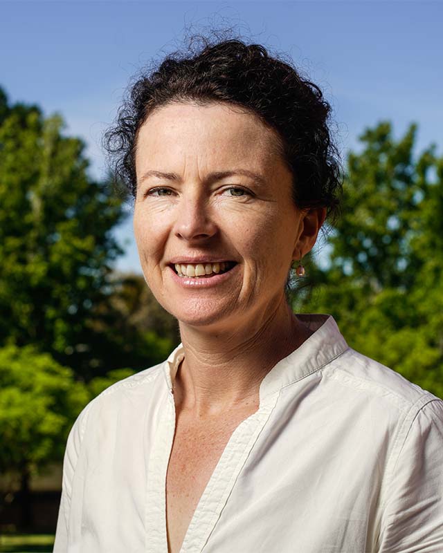 Susie Hester smiling at the camera. She has dark, curly hair, and is wearing a white shirt. There is a blue sky and green trees in the background.