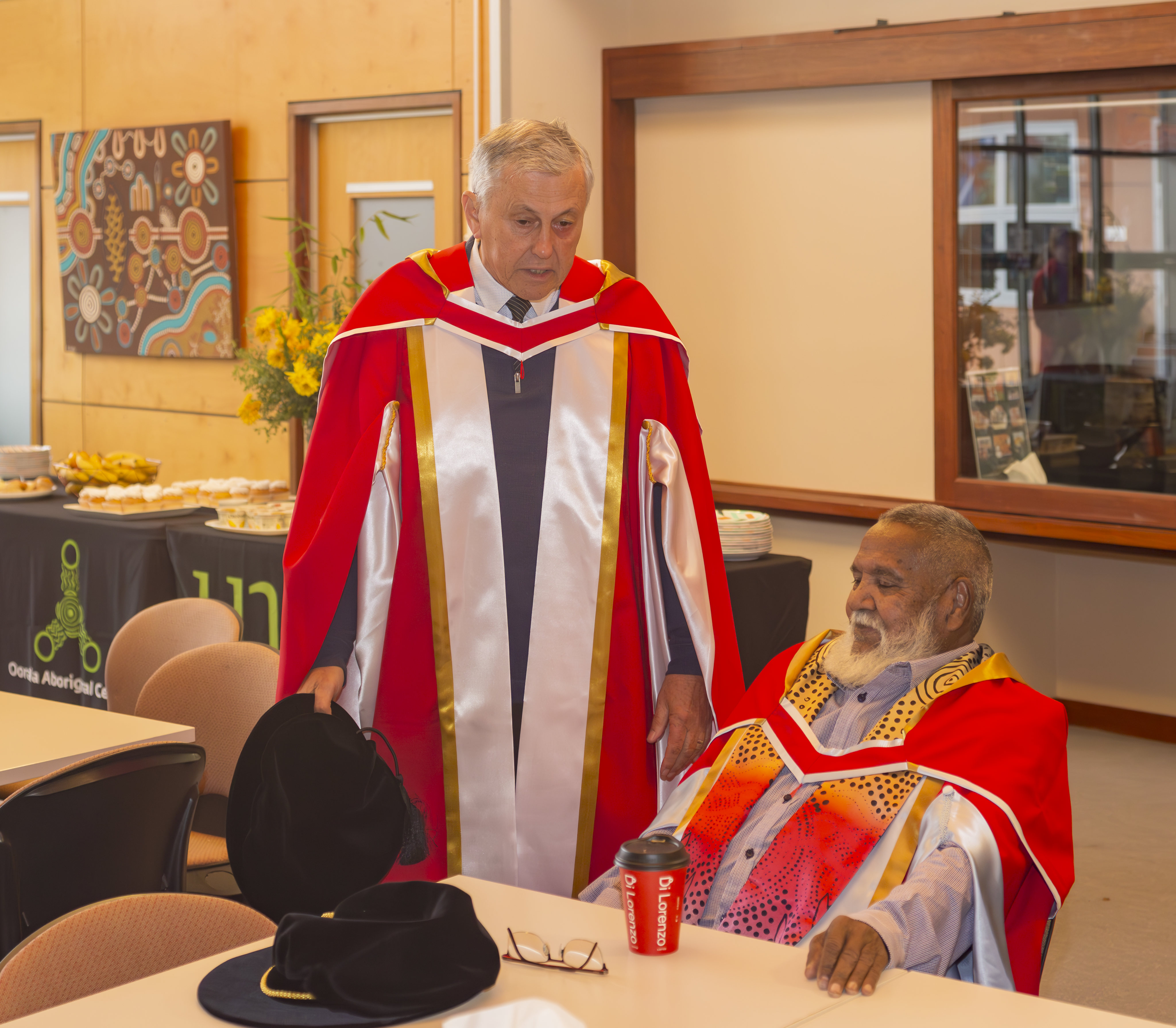 Two elderly Aboriginal men in formal graduation gowns chat with one sitting at a desk.