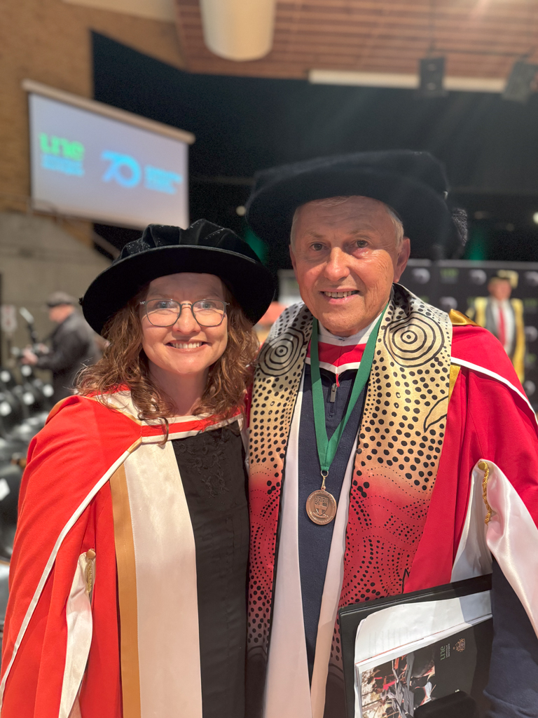 Man and woman in formal graduation gown pose for a photos inside a auditorium. 