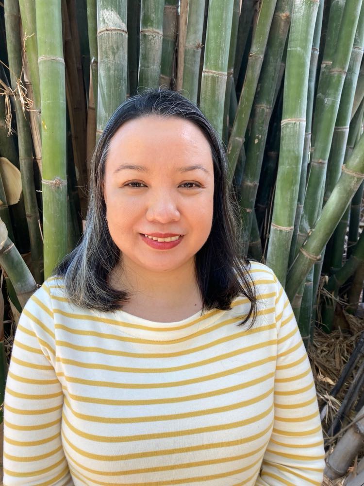 Female academic in white and yellow striped shirt smiles in front of bamboo tree for a photo.