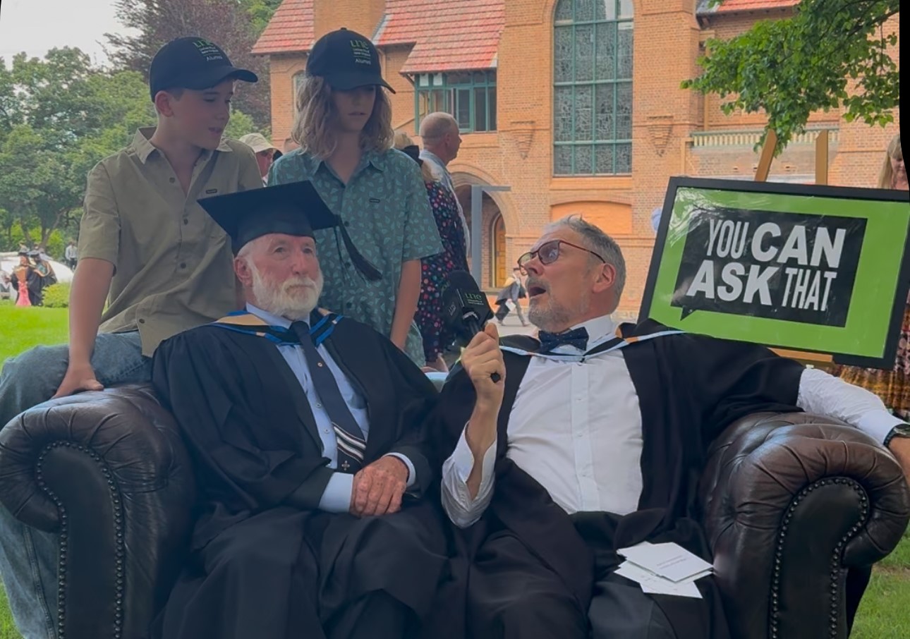 Graduate and his grandchildren sit on a couch with academic for interview on uni lawns at graduation.