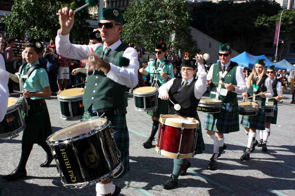 Drummer section of a pipe band in green tartan and hats march through a crowded square. 