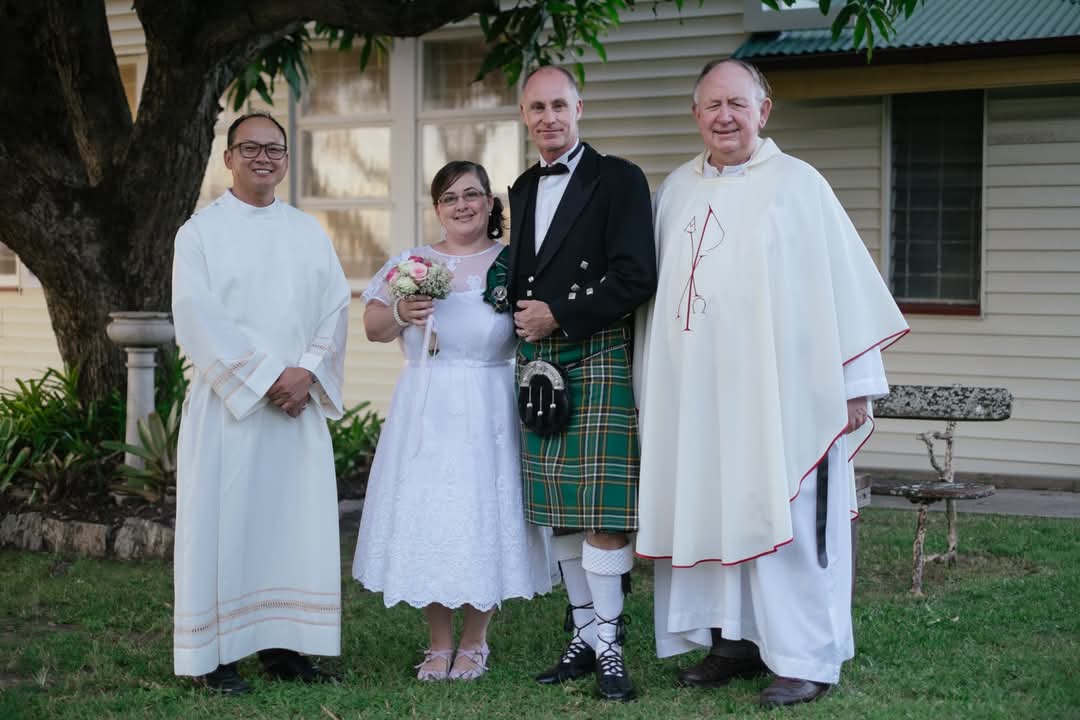 Newly weds in wedding dress and traditional tartan suit stand between two priests and pose for photo.