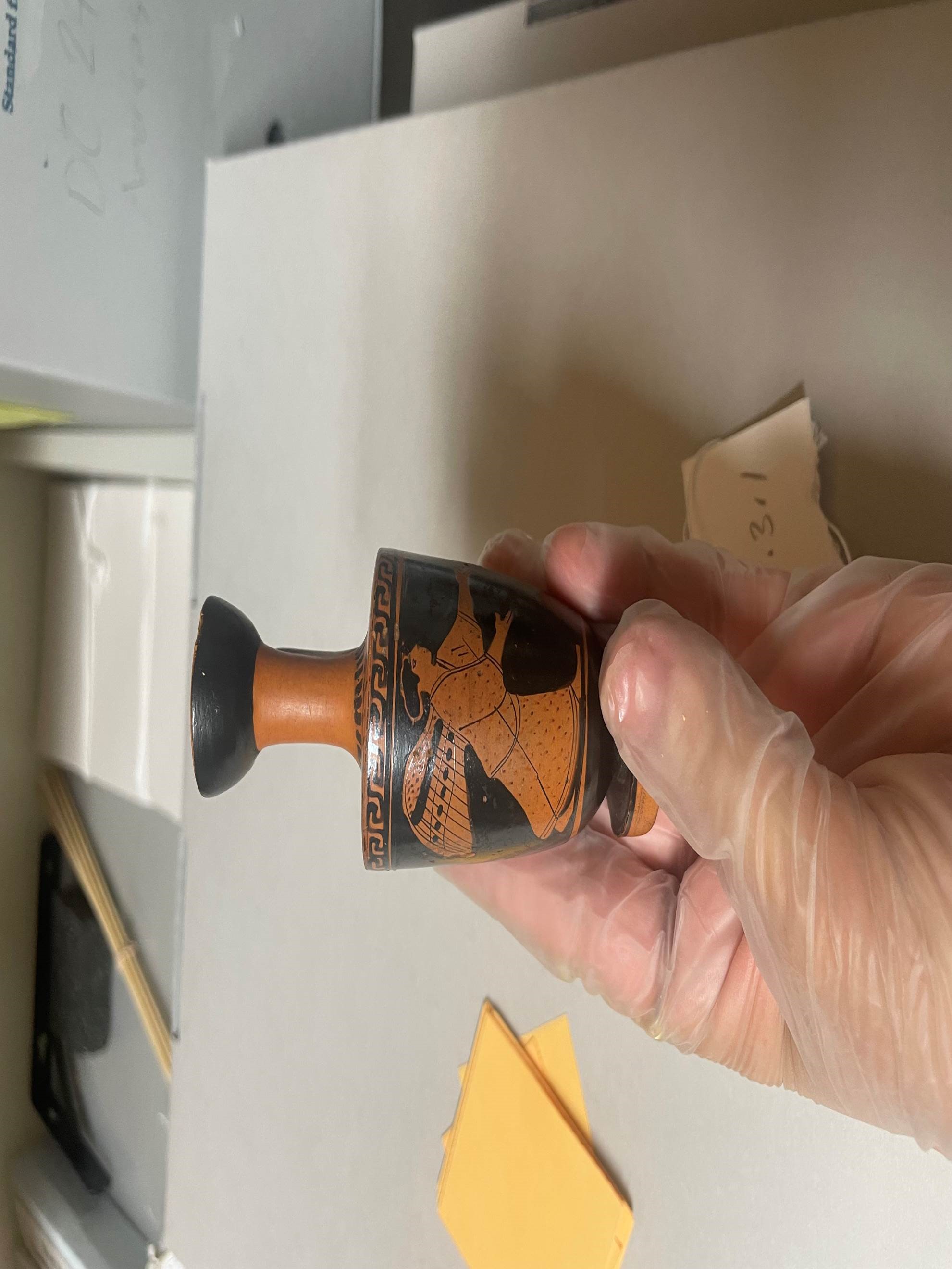 Hand holding a small ancient Greek terracotta vase featuring an image of Nike goddess of victory 