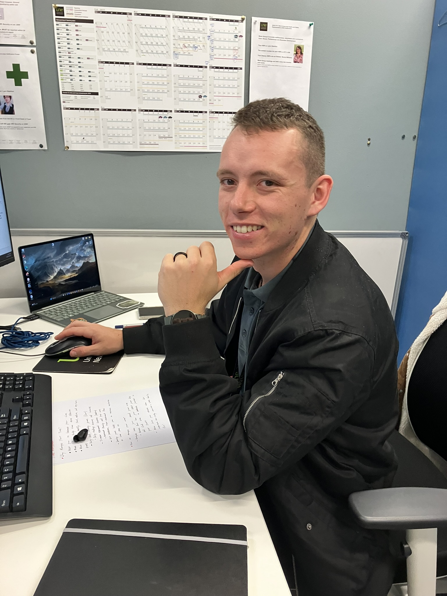 An image of a young man, Cody David, sitting at his work desk in front of his computer, smiling. 