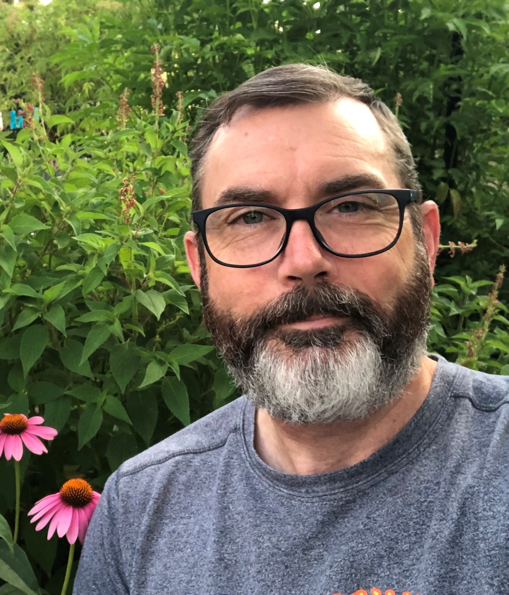 An image of a bearded man surrounded by flowers, smiling at the viewer. 