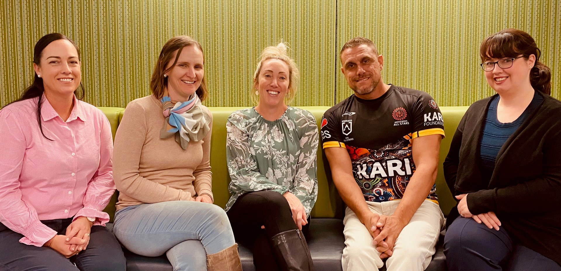 The five UNE Tamworth staff members sit on a green lounge smiling in a welcoming fashion.