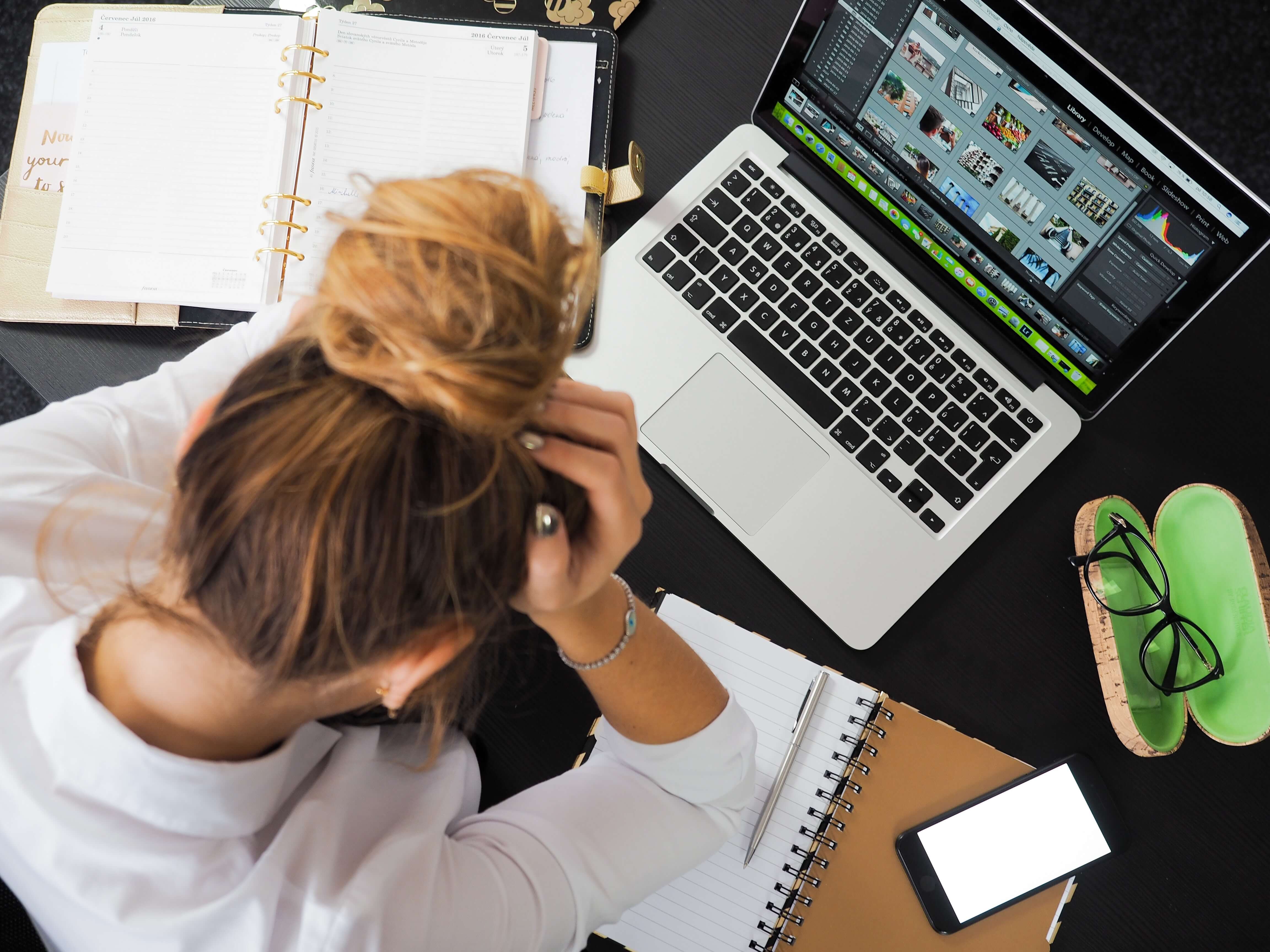 Woman sitting at desk in front of a laptop, glasses case, and notebooks, with her head in her hands