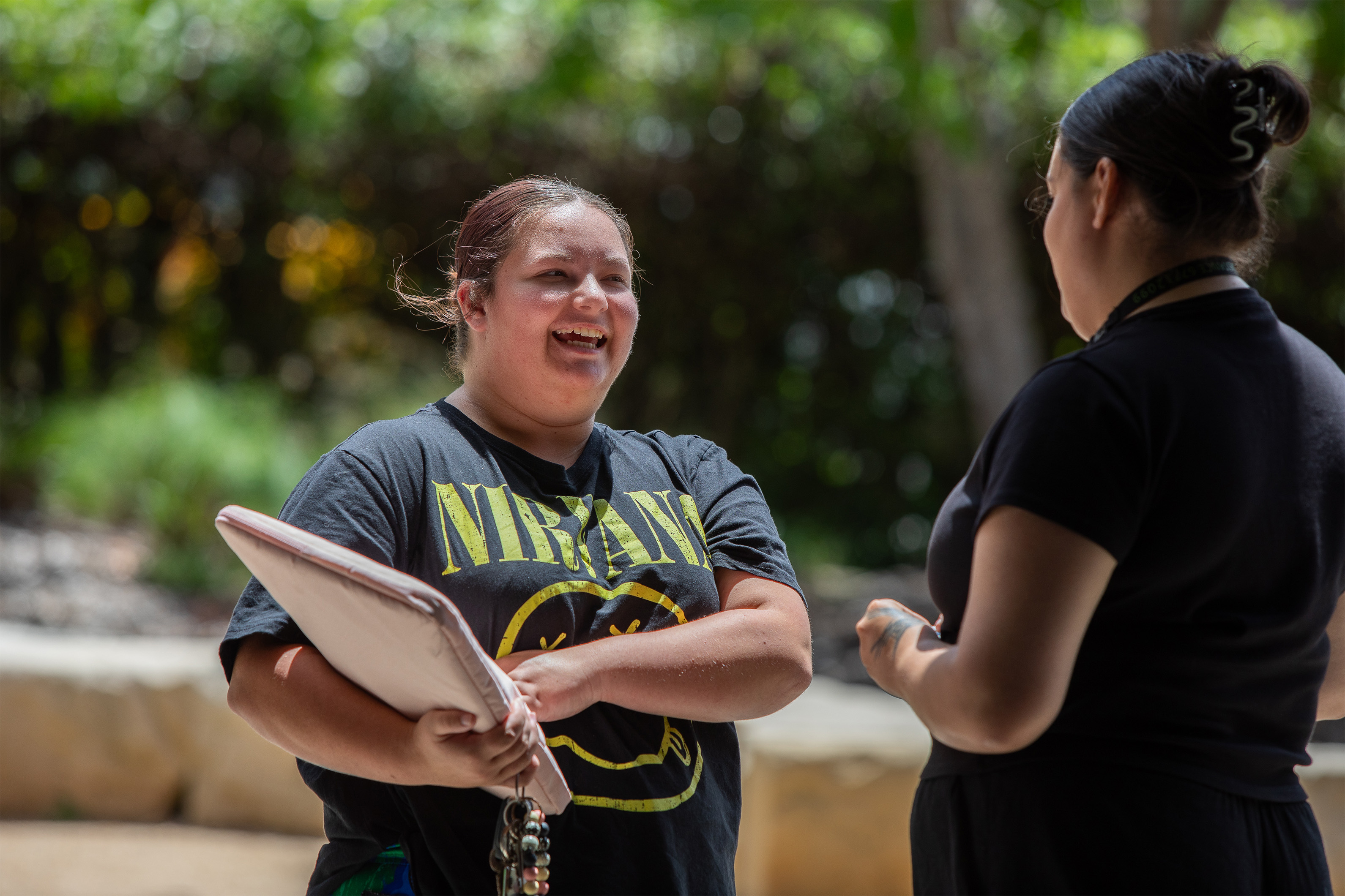 Two women smiling and talking outdoors. One is holding a laptop and wearing a black Nirvana t-shirt, while the other has her back to the camera. They appear to be engaged in a friendly conversation on a sunny day.
