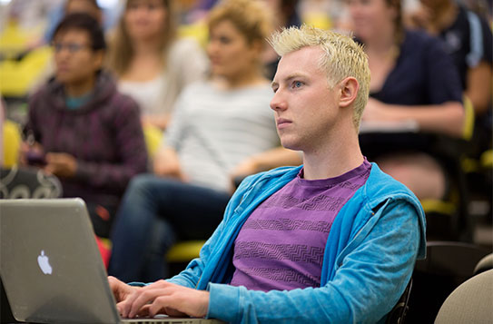 Young male student sitting in lecture theatre with several other students visible in background