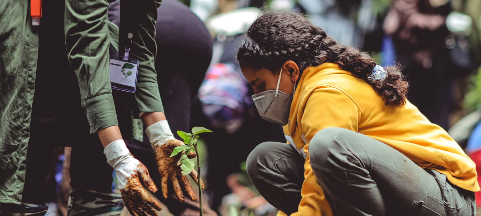 A young girl wearing a mask is crouched down in a field planting a tree, an adult stands beside her helping. 
